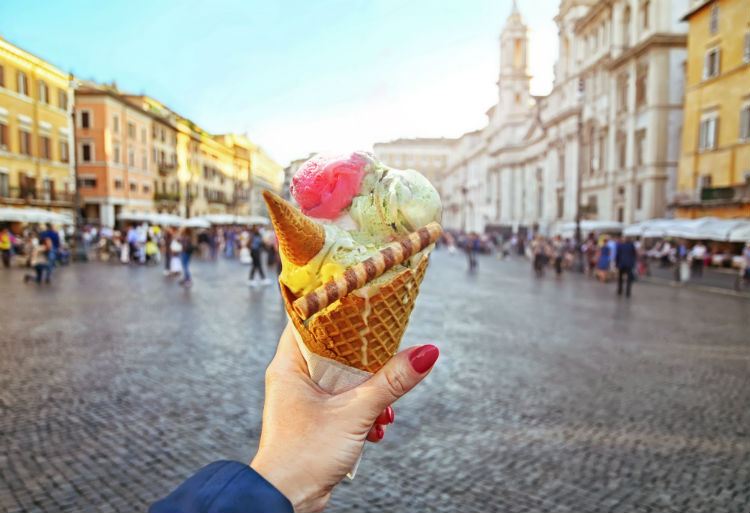 A women's hand with red nails holding up an ice cream cone with an Italian city seen in the background. 