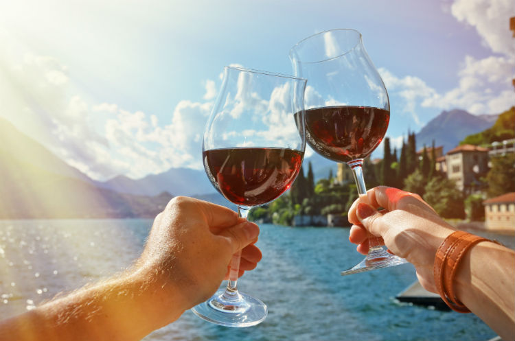 Two hands clinking wine glasses together (which are half full of red wine) with an Italian lake seen in the background. 